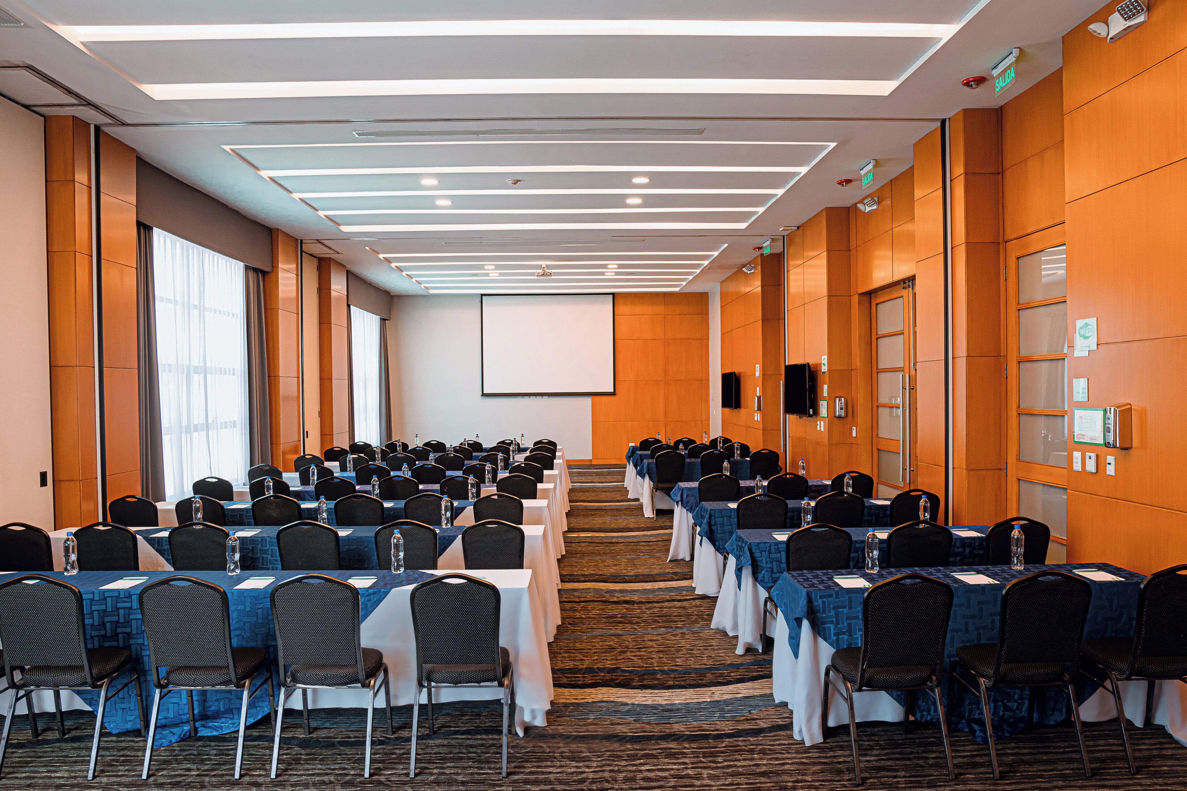 A conference room with rows of tables and chairs, a projector screen, and a carpeted floor.