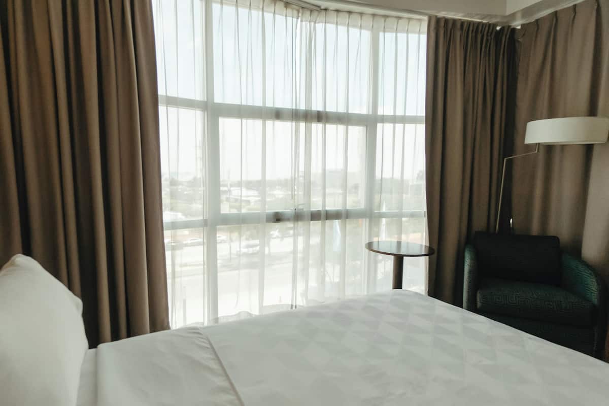 Een hotelkamer met een groot bed, een stoel, een lamp en uitzicht op een stadsgezicht door dunne gordijnen.