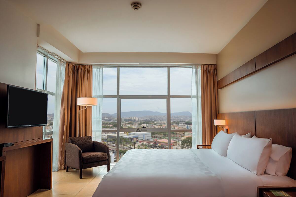 Een hotelkamer met een groot bed, een televisie, een stoel en uitzicht op de stad door grote ramen.