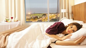 A hotel room with a bed, a nightstand with a lamp, and a view of a cityscape through a large window.