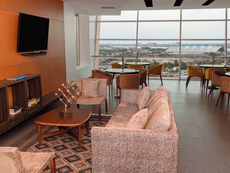 A modern lounge area with a sofa, armchairs, a coffee table, a TV mounted on the wall, and a view of the cityscape through large windows.