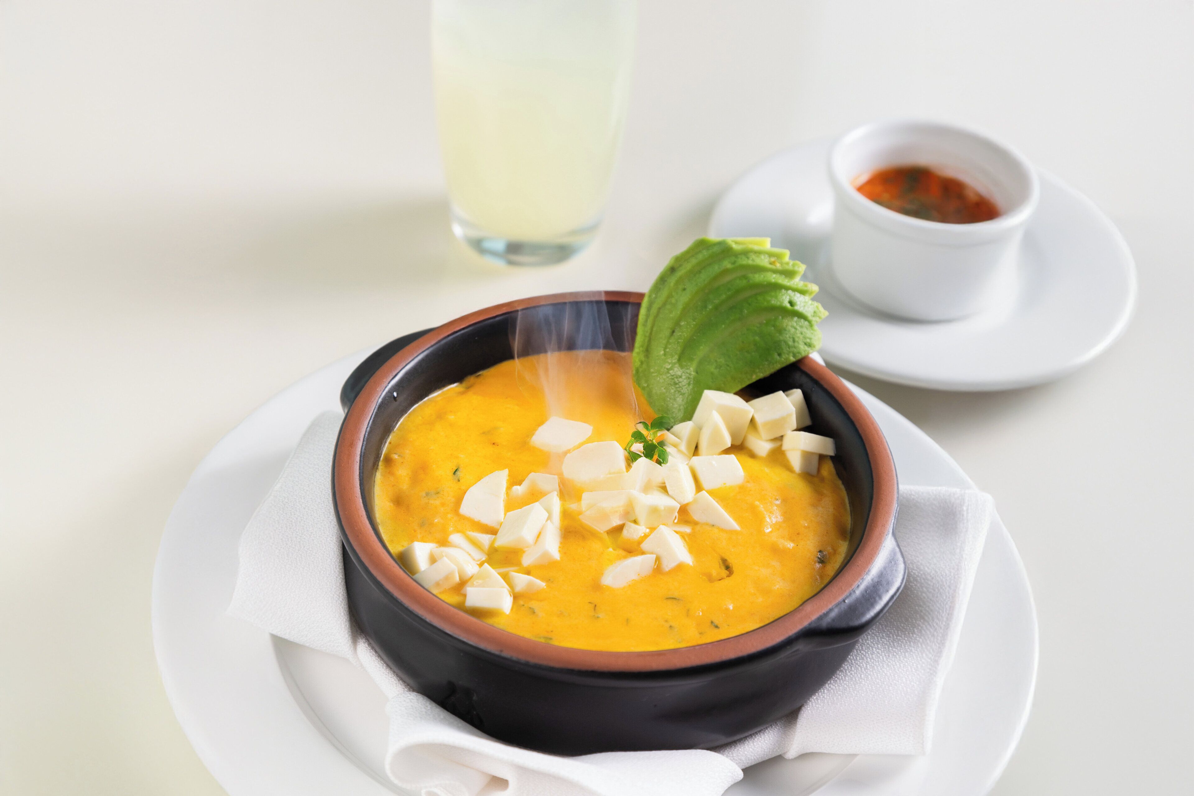 A bowl of soup with a garnish, a small bowl of sauce, and a glass of a clear liquid on a white plate.