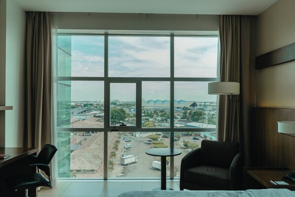 Habitación de hotel con una ventana grande, una cama, una silla, una mesita y una lámpara.
