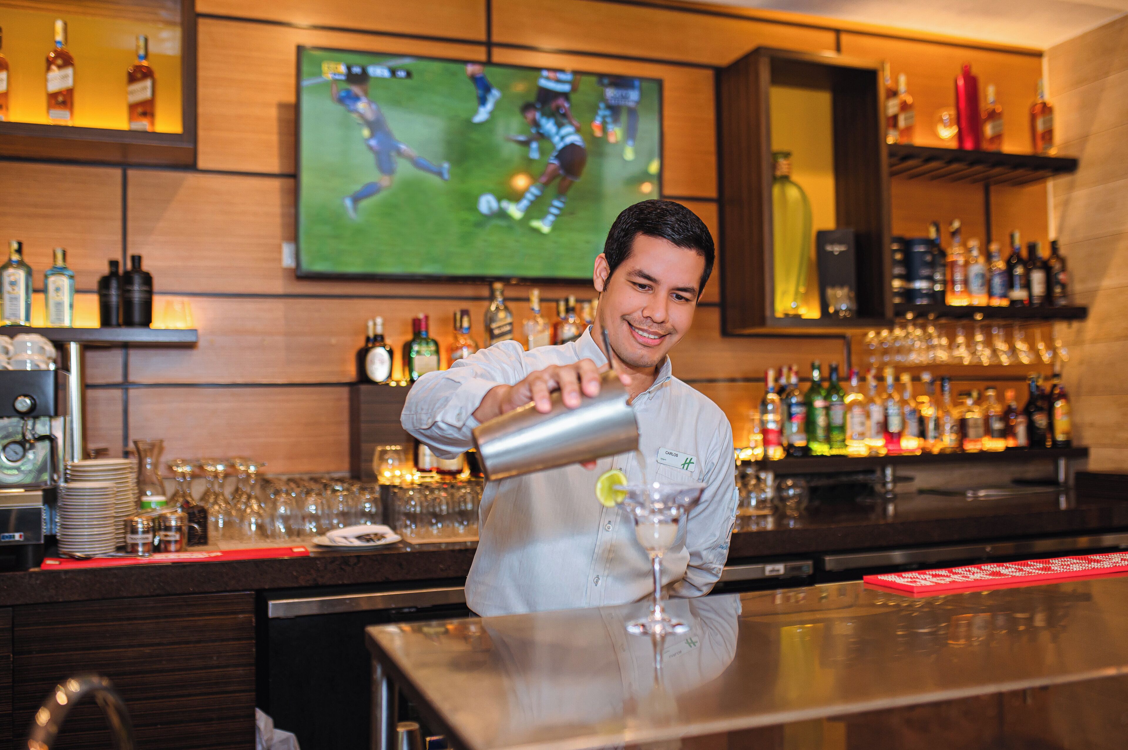 Ein Barkeeper bereitet ein Getränk an einer Bar zu, im Hintergrund läuft ein Sportspiel im Fernsehen. Es sind Regale mit Spirituosen und eine Bar Theke zu sehen.