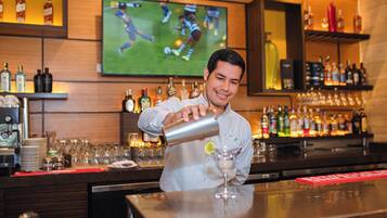 Ein Barkeeper bereitet ein Getränk an einer Bar zu, im Hintergrund läuft ein Sportspiel im Fernsehen. Es sind Regale mit Spirituosen und eine Bar Theke zu sehen.