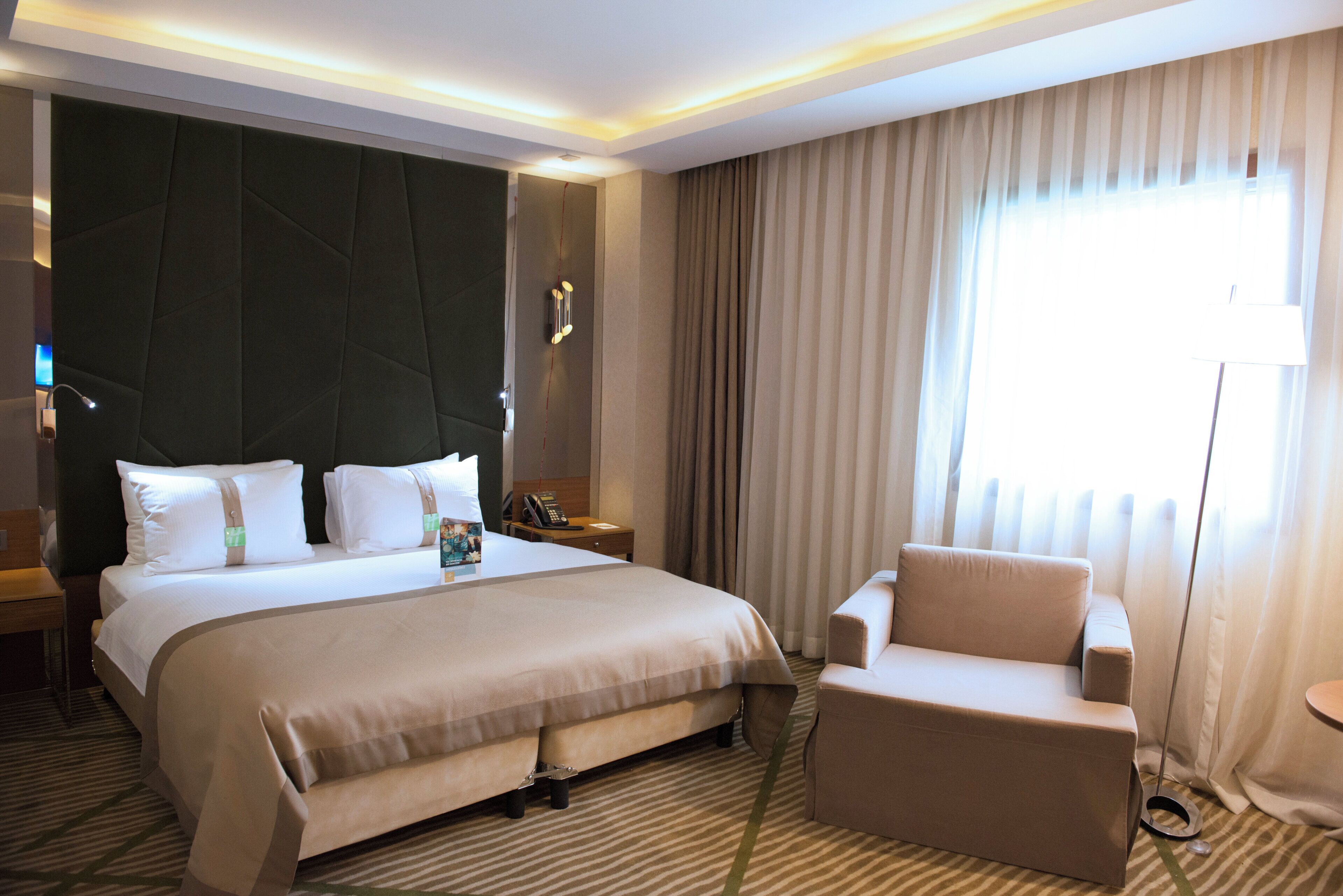 Habitación de hotel con una cama grande, una mesita de noche con teléfono, una silla, un ventanal con cortinas y una alfombra estampada.