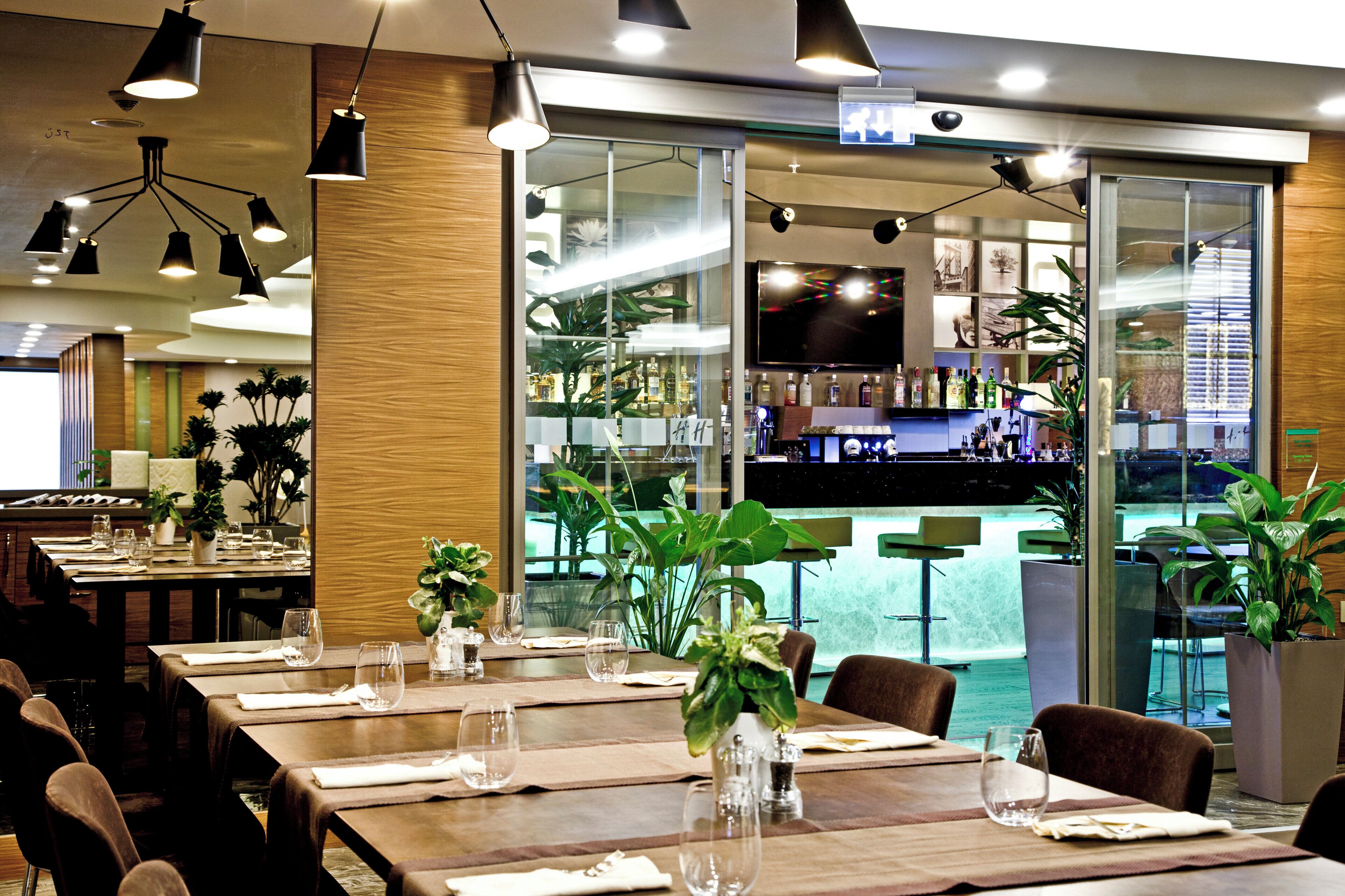 Interno di un ristorante con diversi tavoli, sedie e un'area bar visibile tramite un pannello di vetro.