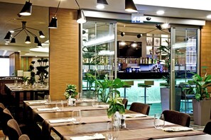 Interior de restaurante con varias mesas, sillas y una zona de bar visible a través de un separador de vidrio.