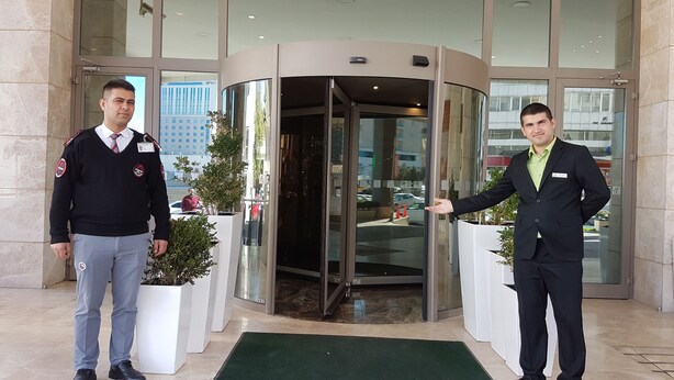 Two men standing in front of a hotel entrance with a revolving door, potted plants, and a glass facade reflecting the cityscape. - Holiday Inn Gaziantep - Sehitkamil by IHG (Gaziantep)
