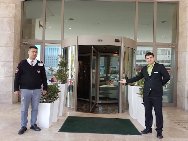 Deux hommes se tiennent devant l’entrée d’un hôtel, qui est dotée d’une porte tournante, de plantes en pot et d’une façade en verre qui reflète le paysage urbain.