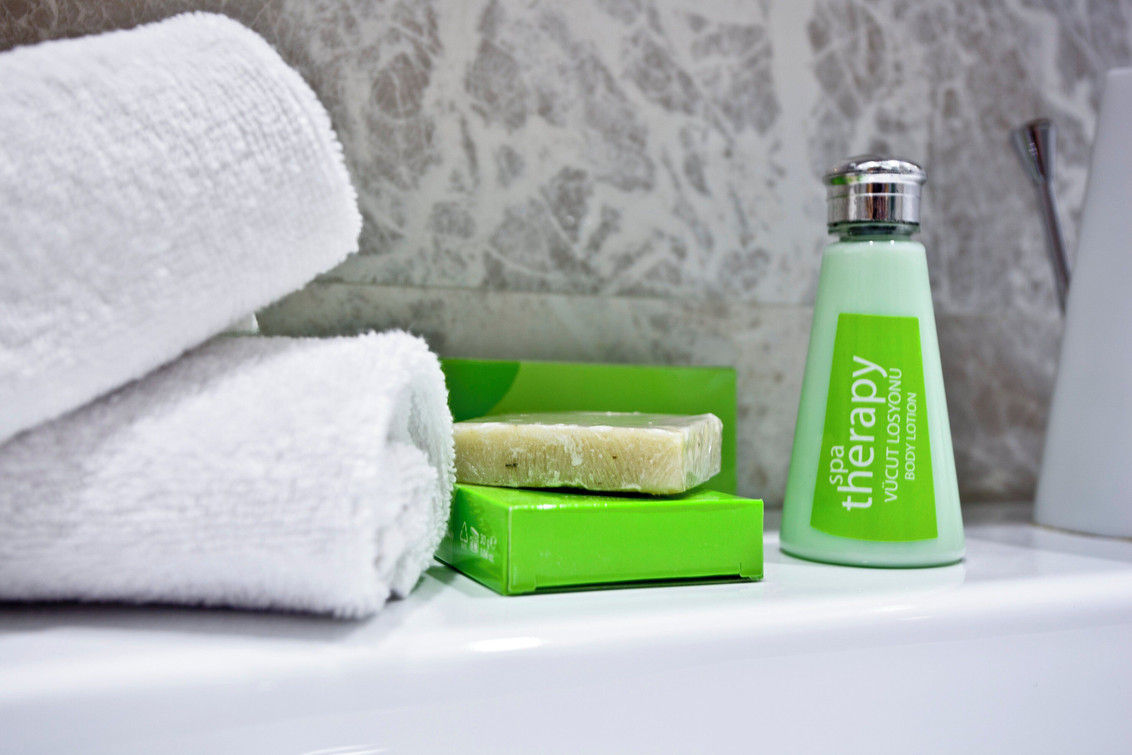 A bathroom with a white towel, a green soap bar, and a green bottle of spa therapy body lotion.