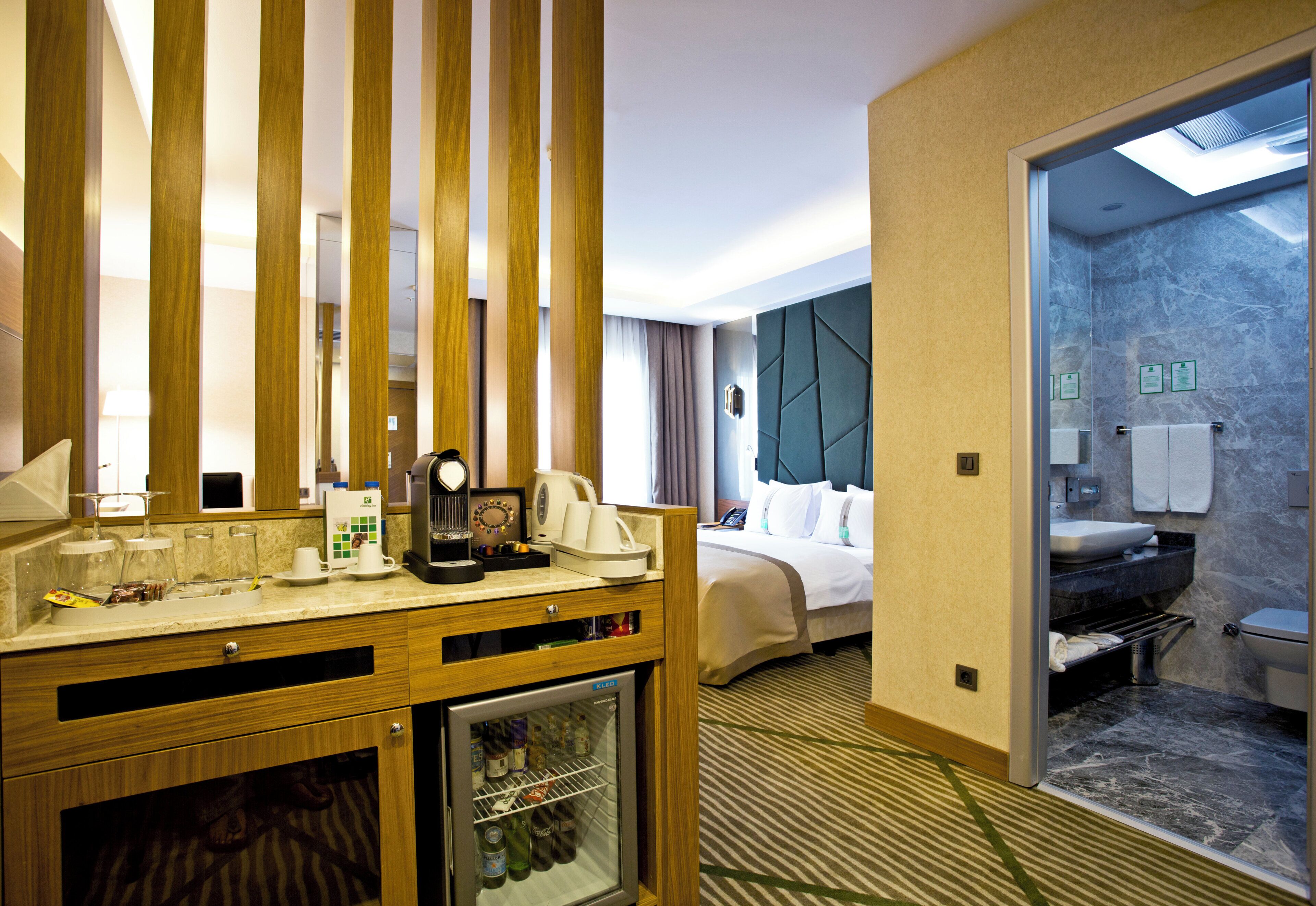 Une chambre d’hôtel avec un minibar, un lit, une salle de bain avec un mur en marbre et une vue sur l’extérieur, visible à travers une porte.