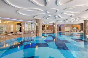 Uma grande piscina coberta com piso ladrilhado e teto com desenhos circulares. Há cadeiras de descanso e uma área de recepção à vista.
