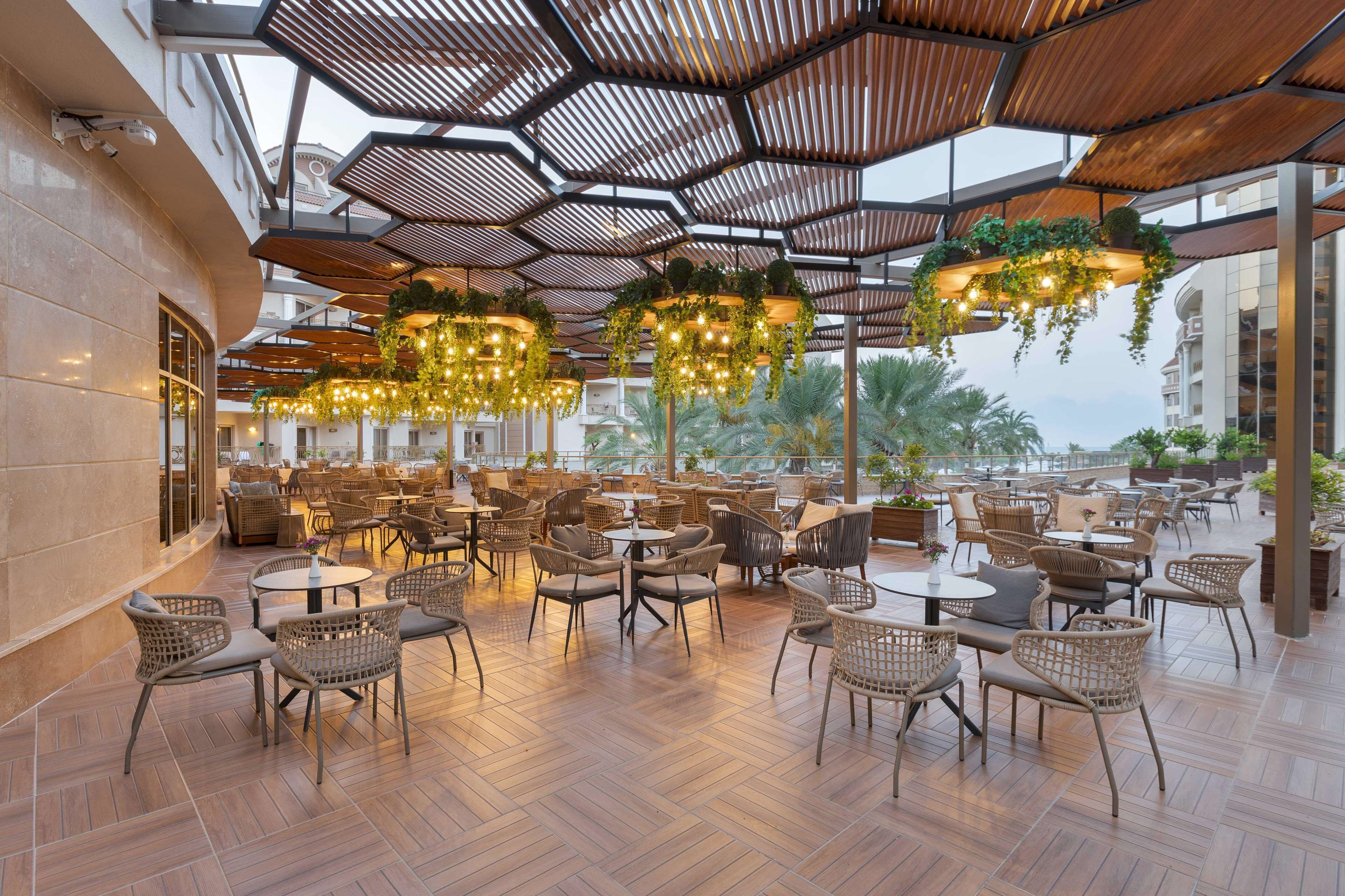 Un café al aire libre espacioso con paneles de techo de madera hexagonal, mobiliario de mimbre y plantas colgantes.