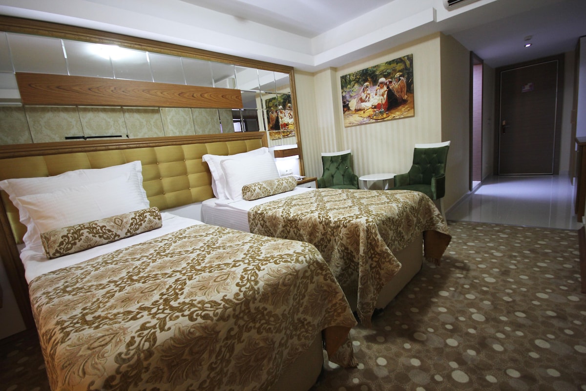 A hotel room with two beds, a large mirror, a green chair, and a painting on the wall.