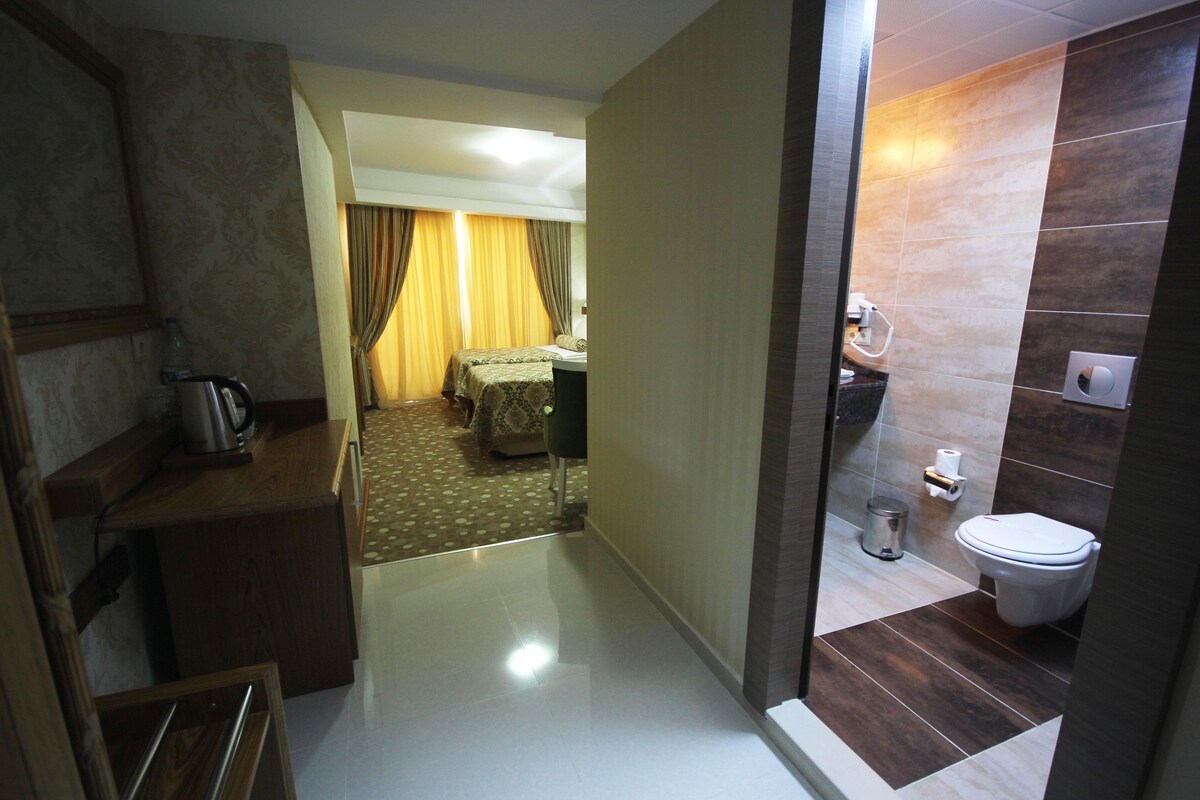 A hotel room with a kitchenette, a bed with a patterned bedspread, a bathroom with a toilet and sink, and a hallway leading to another room.