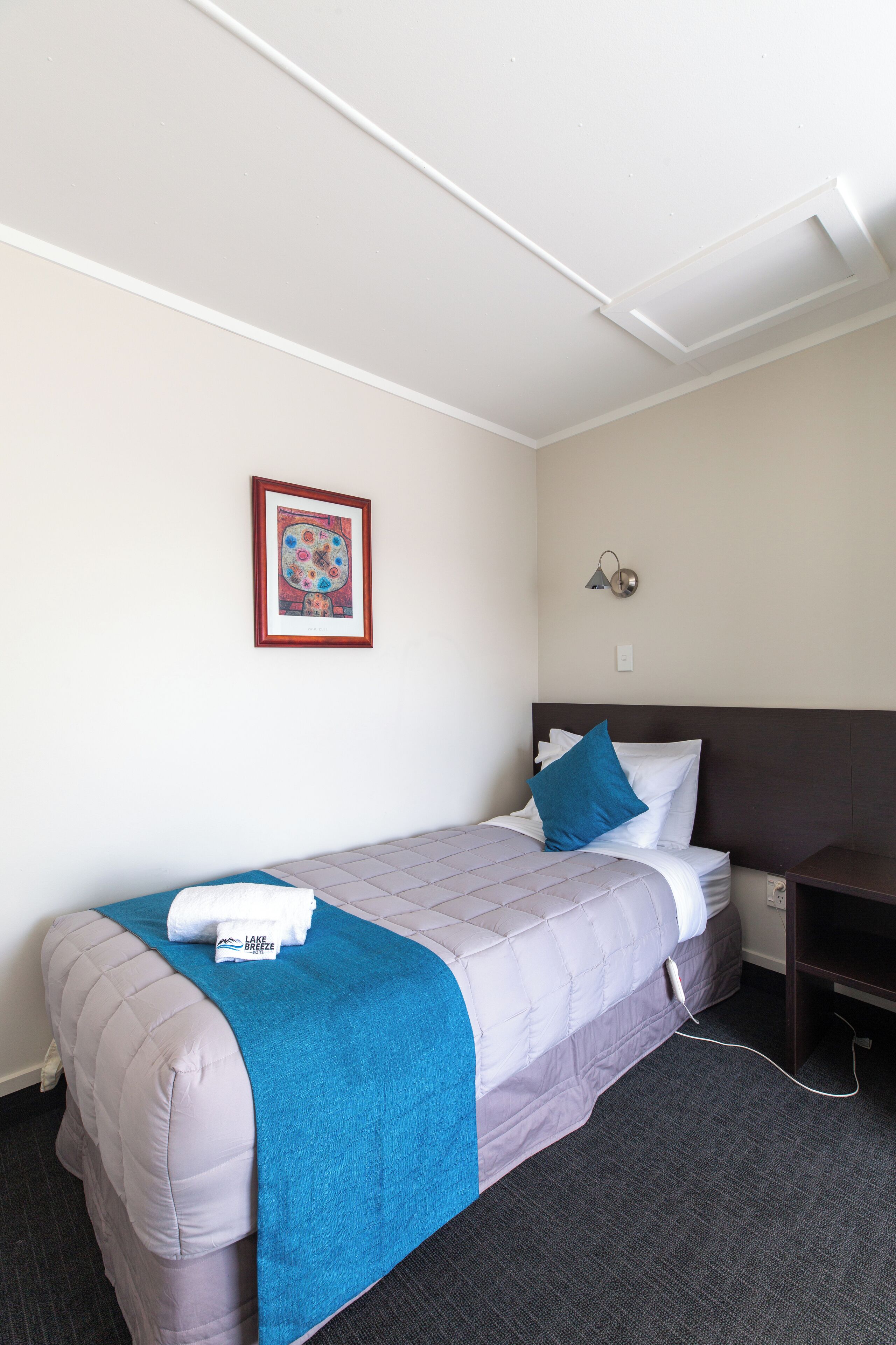 A neatly made bed with a blue blanket and pillows, a framed picture on the wall, and a bedside table with a lamp.