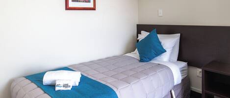 A neatly made bed with a blue blanket and pillows, a framed picture on the wall, and a bedside table with a lamp.
