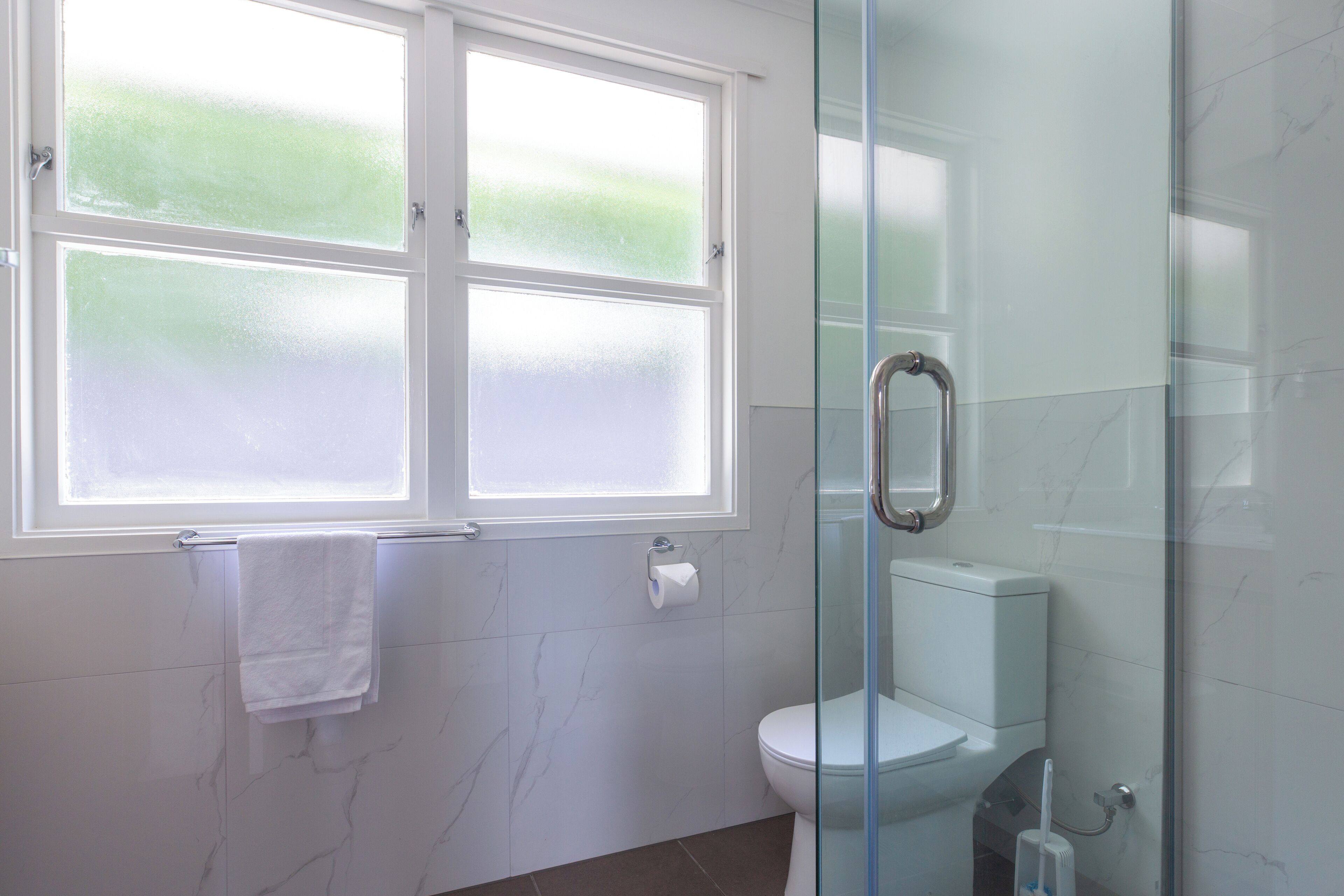 A bathroom with a toilet, a glass shower enclosure, a window with frosted glass, and a towel hanging on the wall.