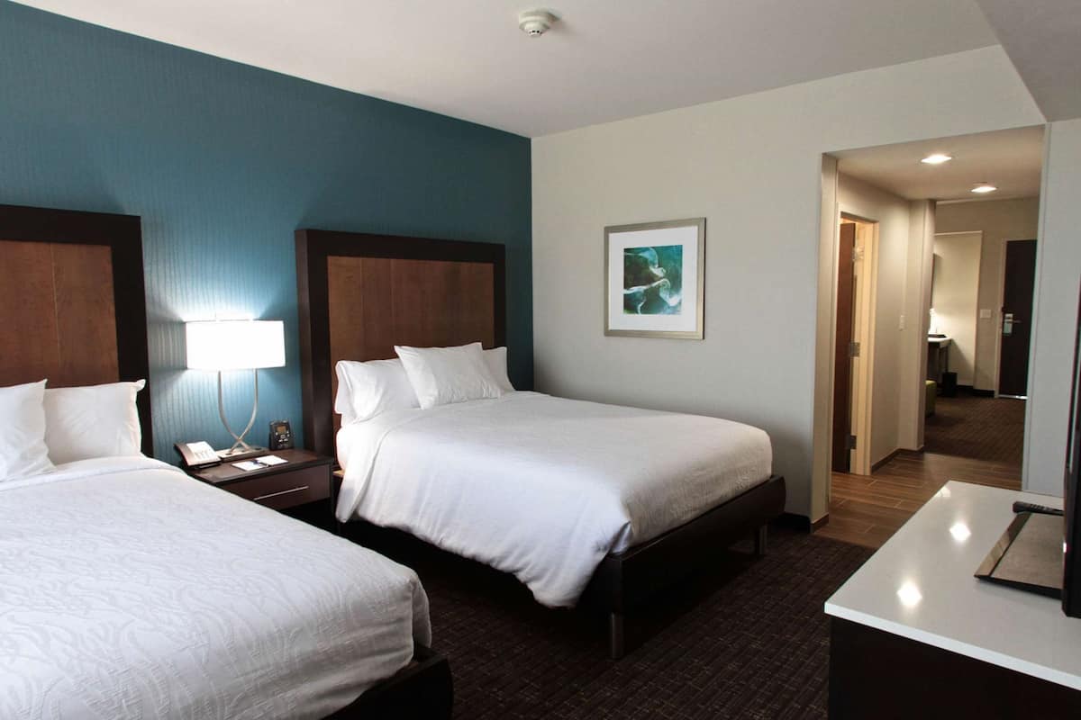 A hotel room with two beds, a desk, a nightstand, a lamp, and a framed picture on the wall.