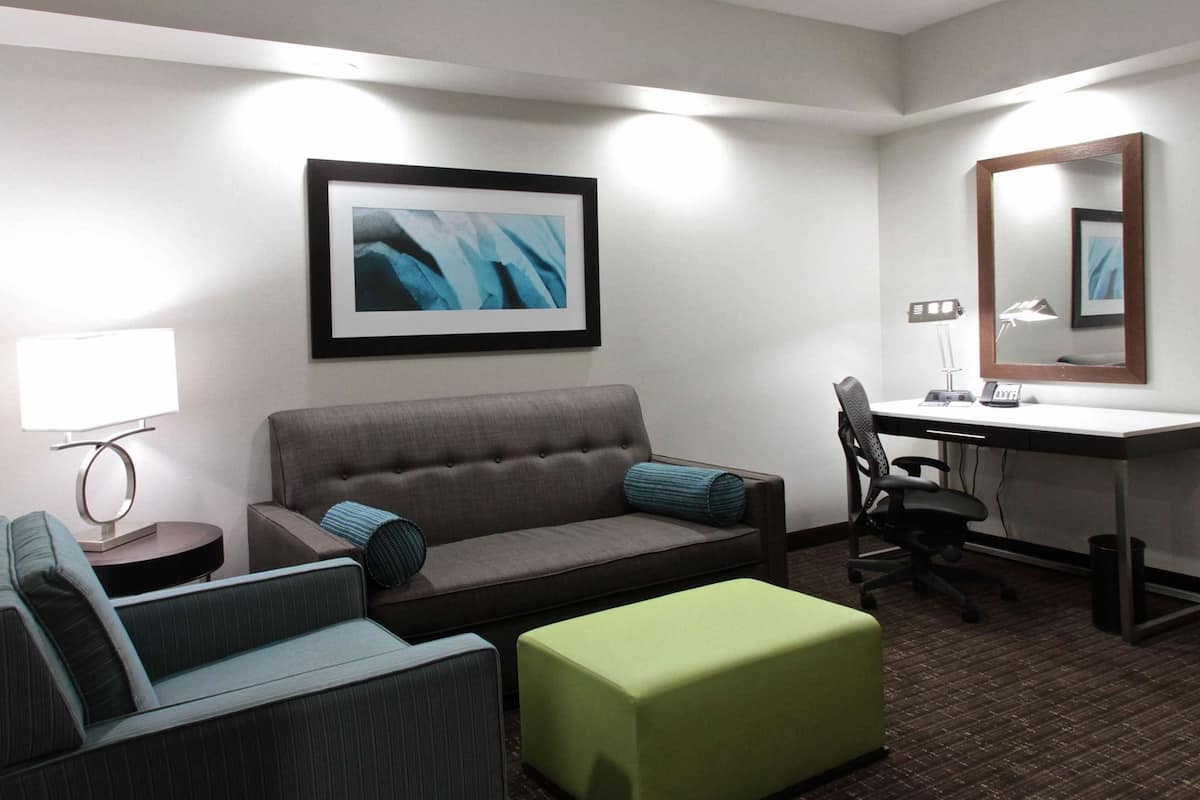 A hotel room with a sofa, a green ottoman, a desk with a chair, a lamp, and a framed abstract painting.