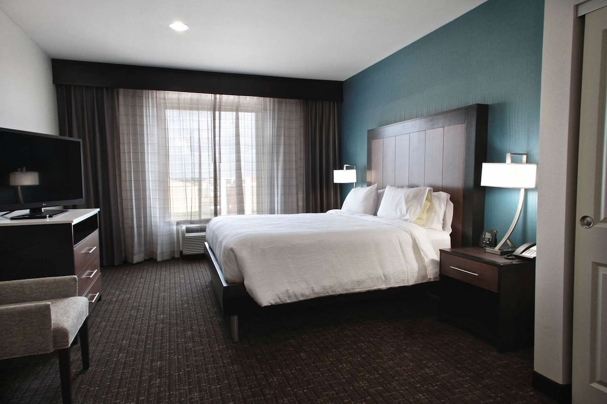 A hotel room with a bed, a desk, a chair, a TV, a bedside table with a lamp, and a window with curtains.