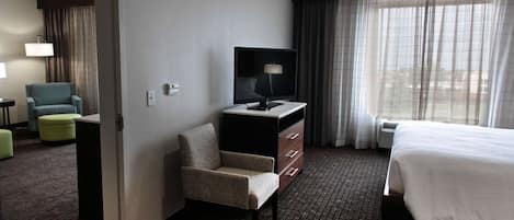 A hotel room with a bed, a desk with a TV, a chair, and a large window with curtains.