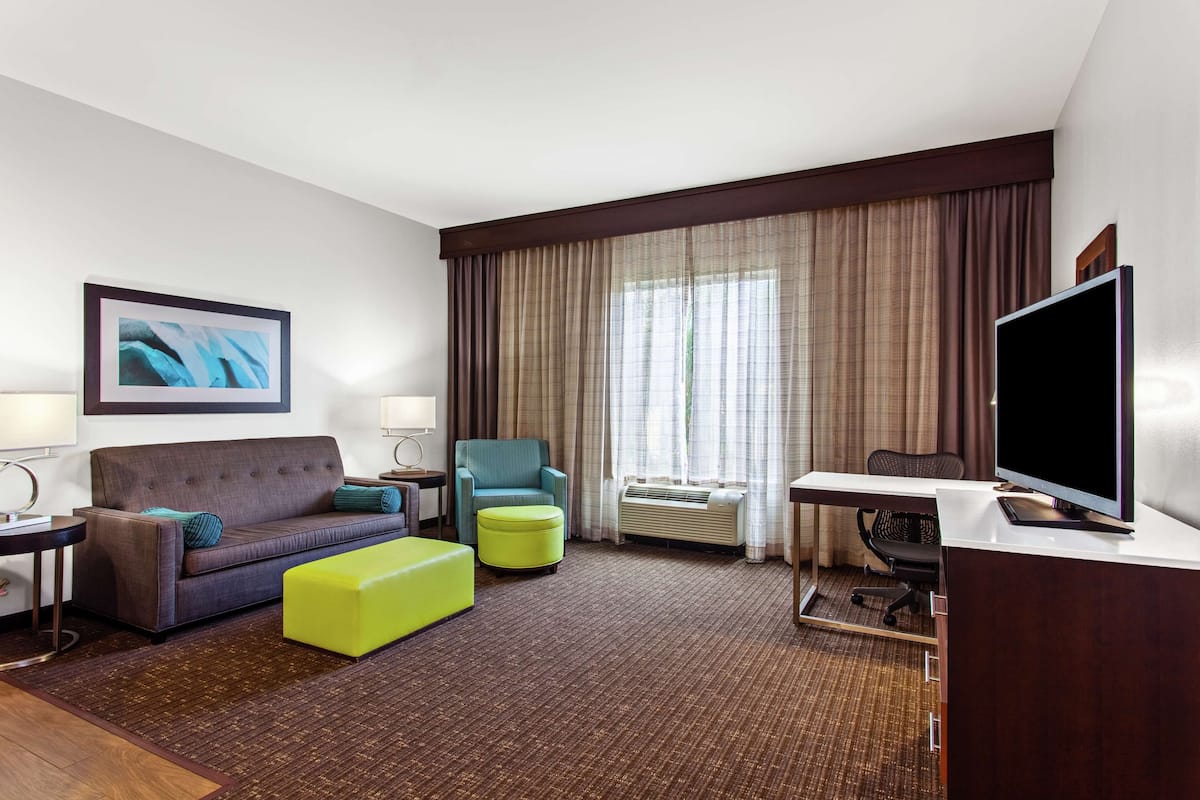 A hotel room with a sofa, armchair, ottoman, desk, television, and a large window with curtains.