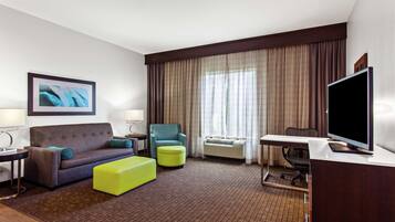 Habitación de hotel con sofá, sillón, reposapiés, escritorio, televisor y un amplio ventanal con cortinas.