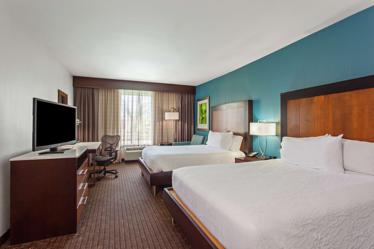 A hotel room with two beds, a desk with a flat-screen TV, a chair, and a window with curtains.