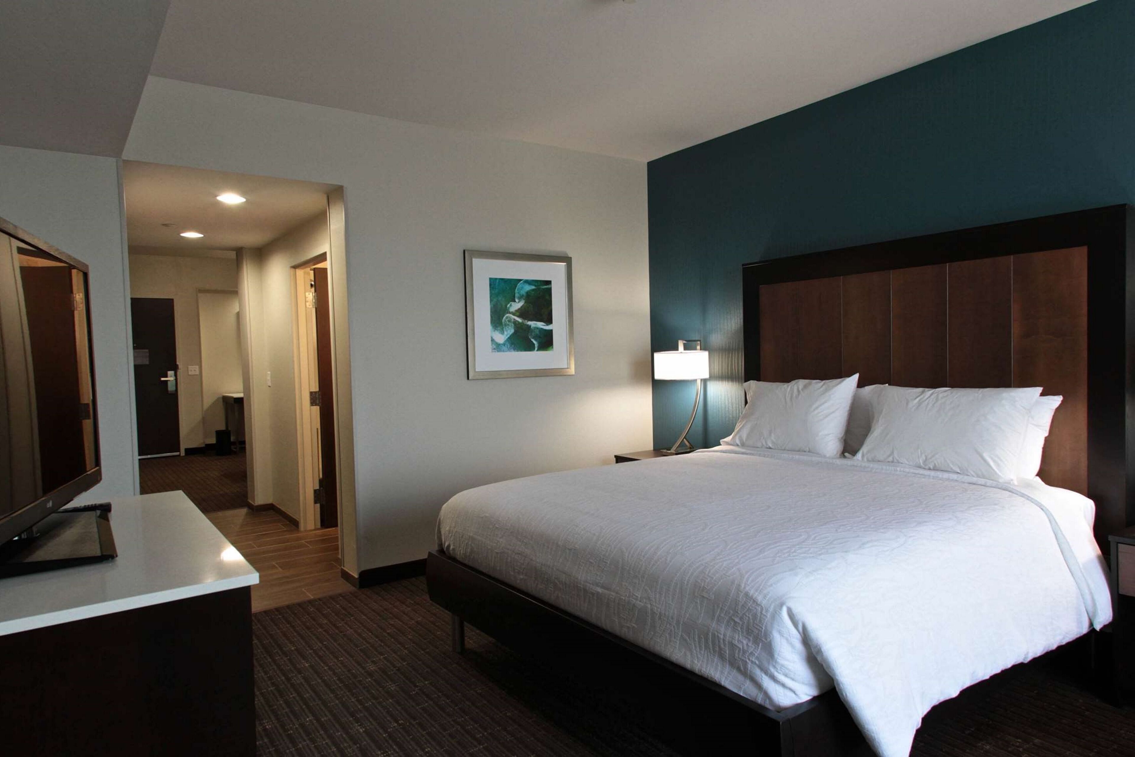 Quarto de hotel com uma cama grande, uma escrivaninha com televisão de tela plana, um abajur e um quadro na parede.