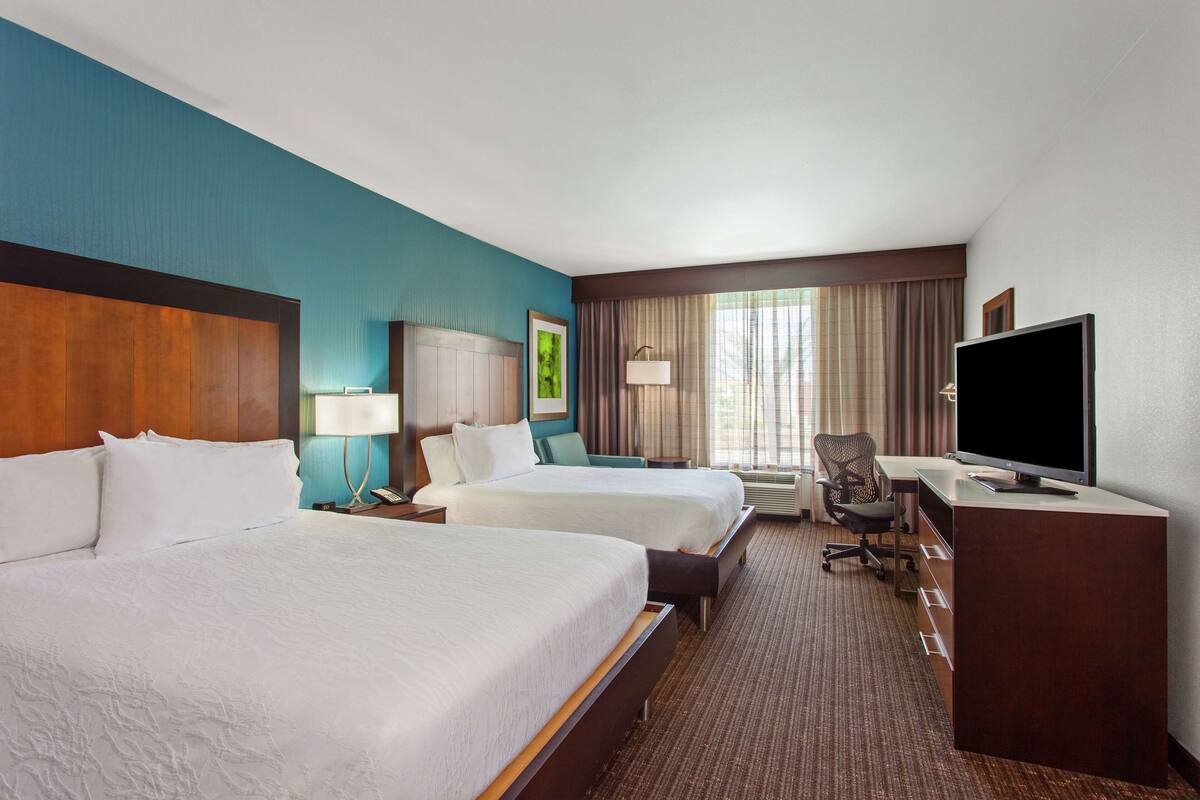 A hotel room with two beds, a desk, a TV, and a window with curtains.