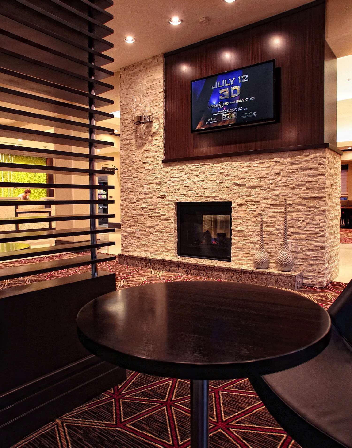 A modern interior with a stone wall, a flat-screen TV displaying 'JULY 12 3D', a fireplace, and a round table with chairs.
