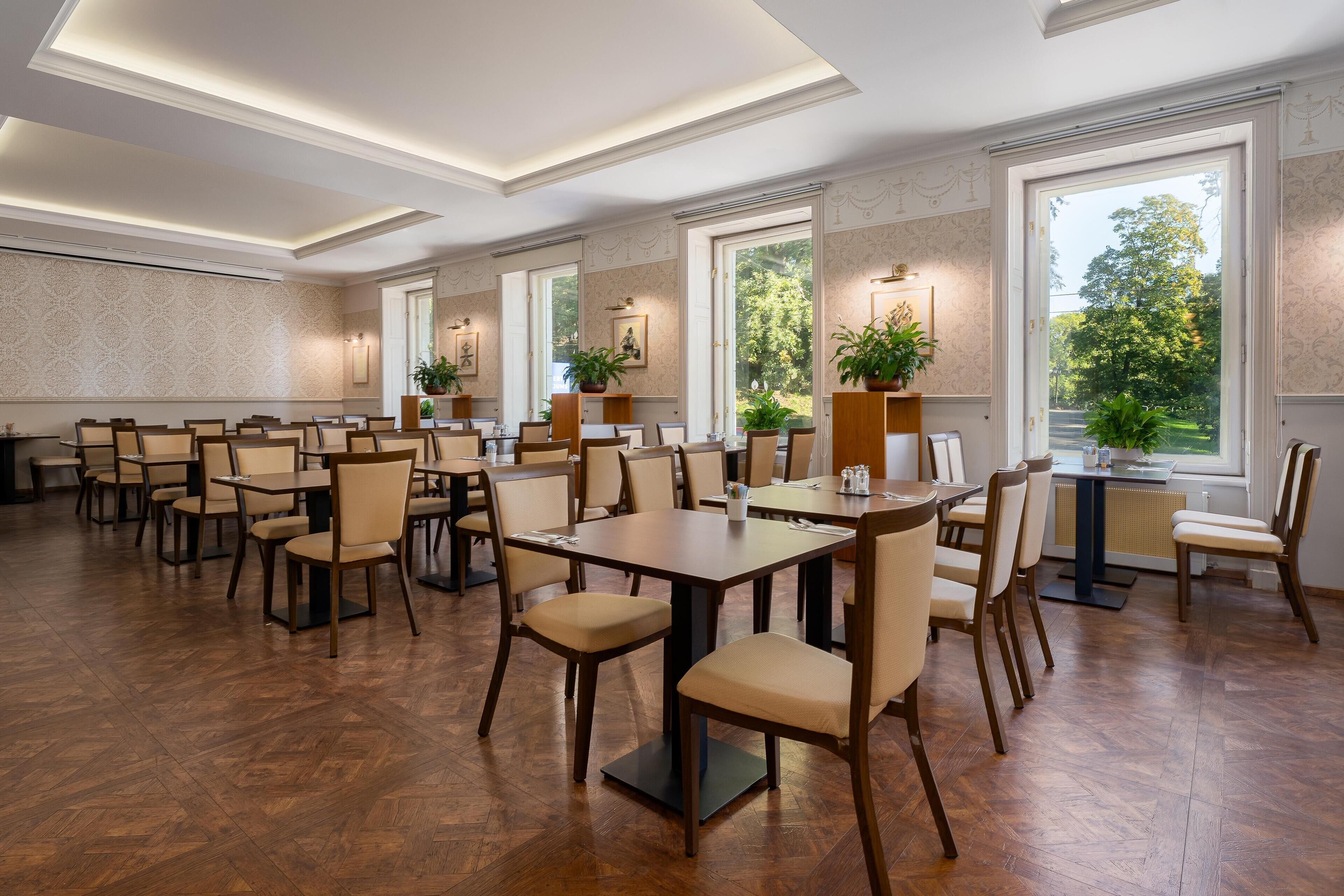 A spacious dining area with multiple tables and chairs, large windows offering a view of the outdoors, and a ceiling with recessed lighting.