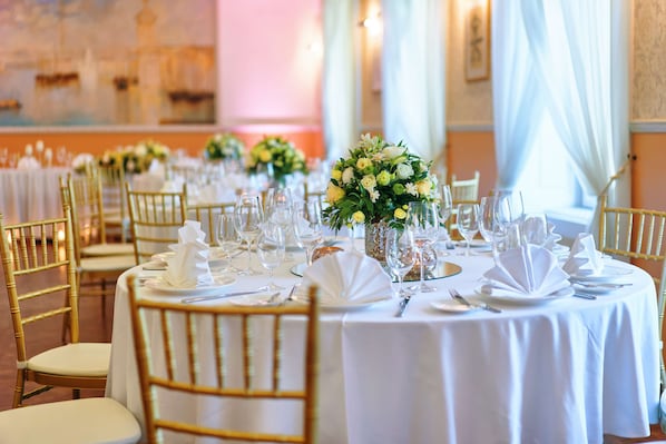 Ambiente formal para comedor con mesas redondas, sillas doradas, manteles blancos y centros de mesa con flores.