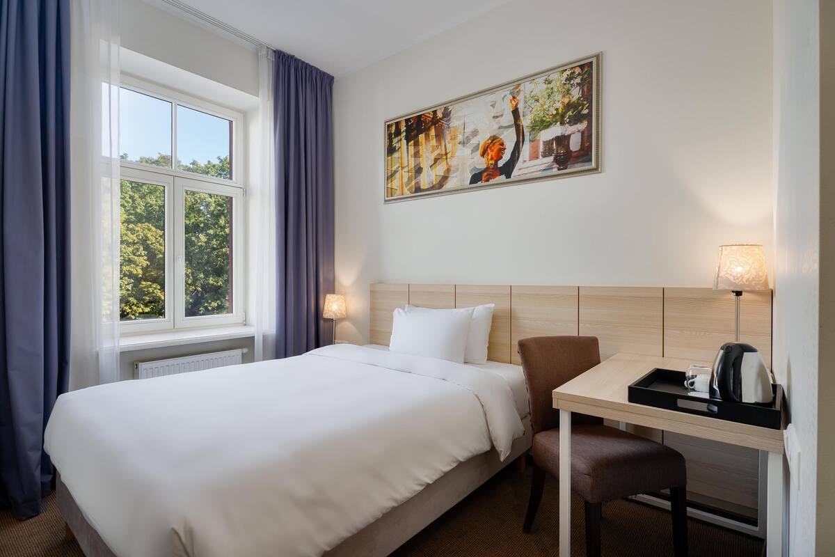 A hotel room with a large bed, a desk with a chair, a painting on the wall, and a window with curtains.