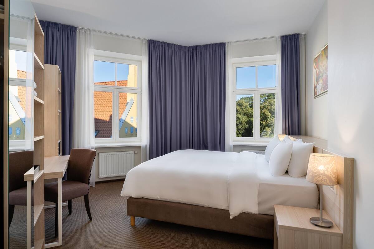 A hotel room with a bed, a desk, a chair, and a lamp.