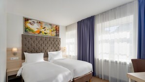A hotel room with two beds, a tufted headboard, a painting above the beds, a window with sheer curtains, and a table with a chair. - Radisson Hotel Old Town Riga (Riga)