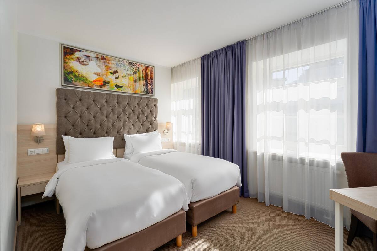 A hotel room with two beds, a tufted headboard, a painting above the beds, a window with sheer curtains, and a table with a chair.