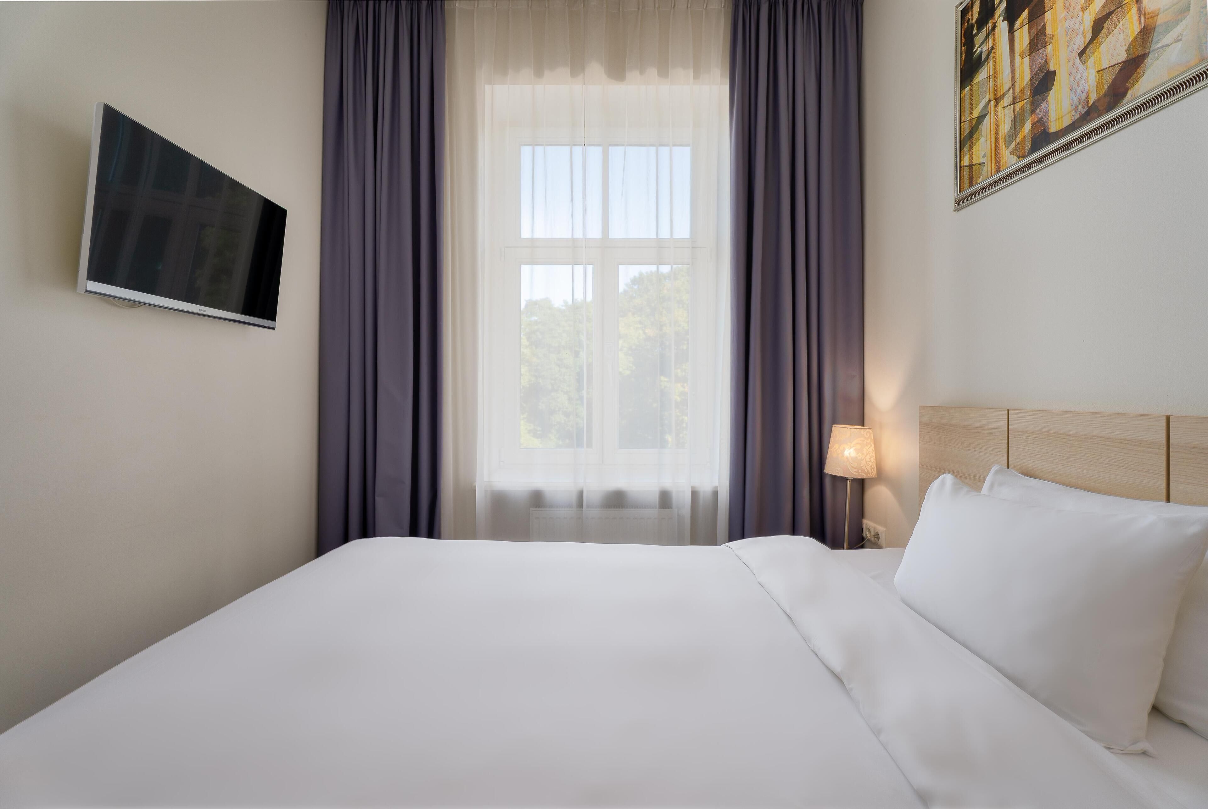A hotel room with a bed, a flat-screen TV mounted on the wall, a window with curtains, and a painting on the wall.