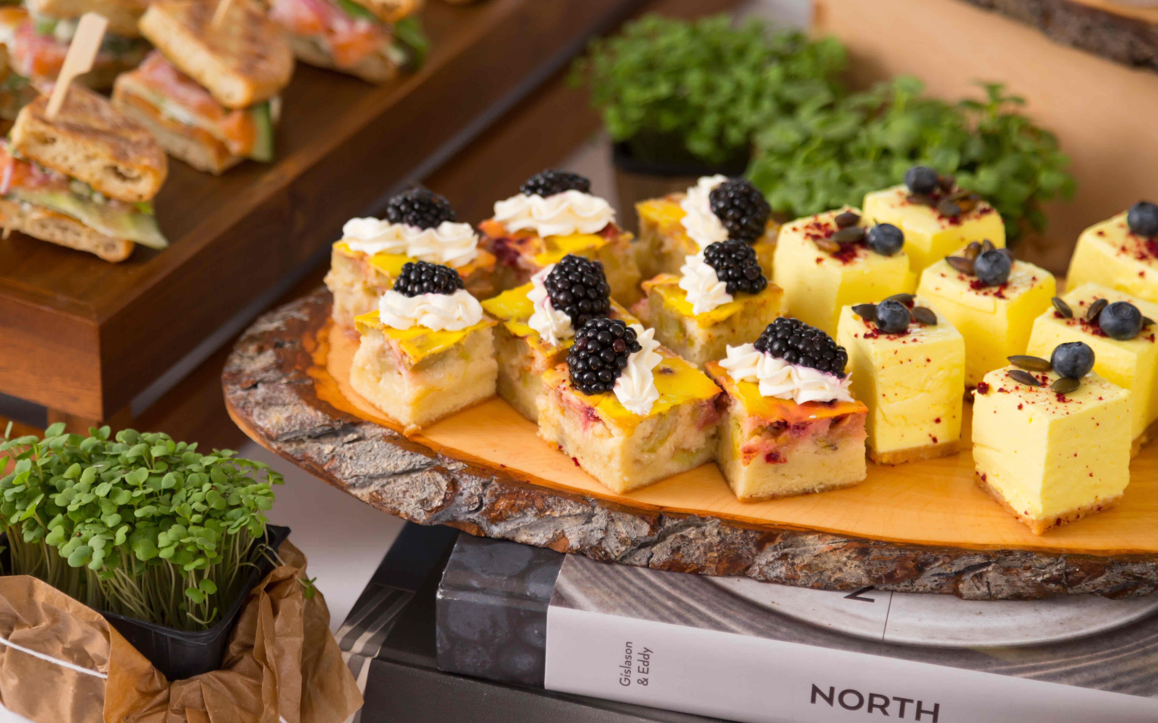 A variety of desserts on a wooden board, including squares topped with blackberries and whipped cream.