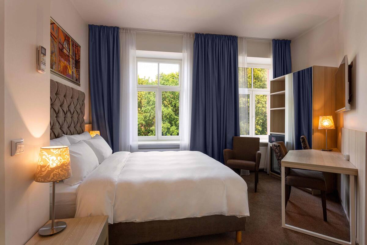 A hotel room with a large bed, a desk, a chair, a TV, and a window with curtains.