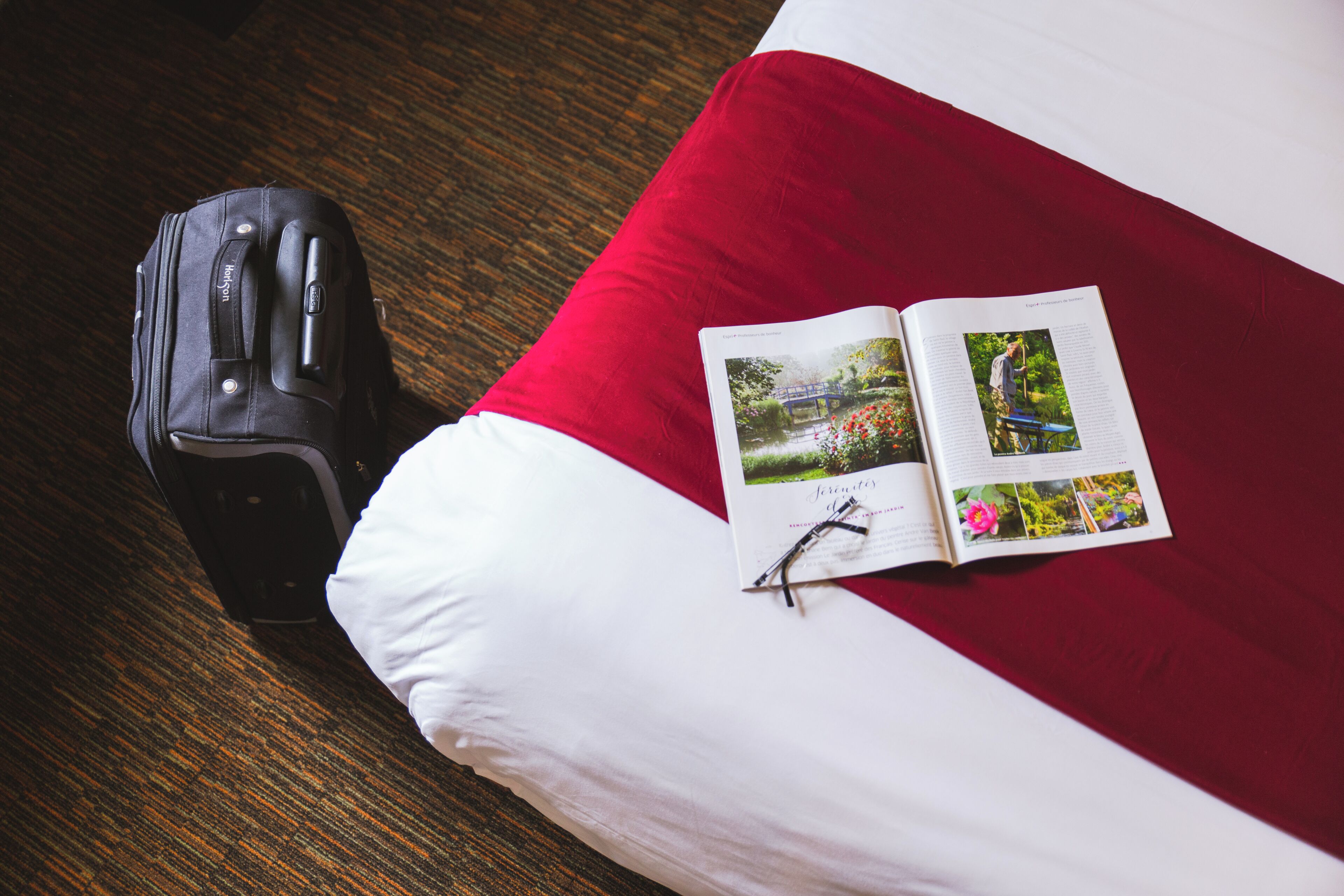 Un lit avec un oreiller rouge et blanc, une valise noire et un magazine ouvert avec des photos.