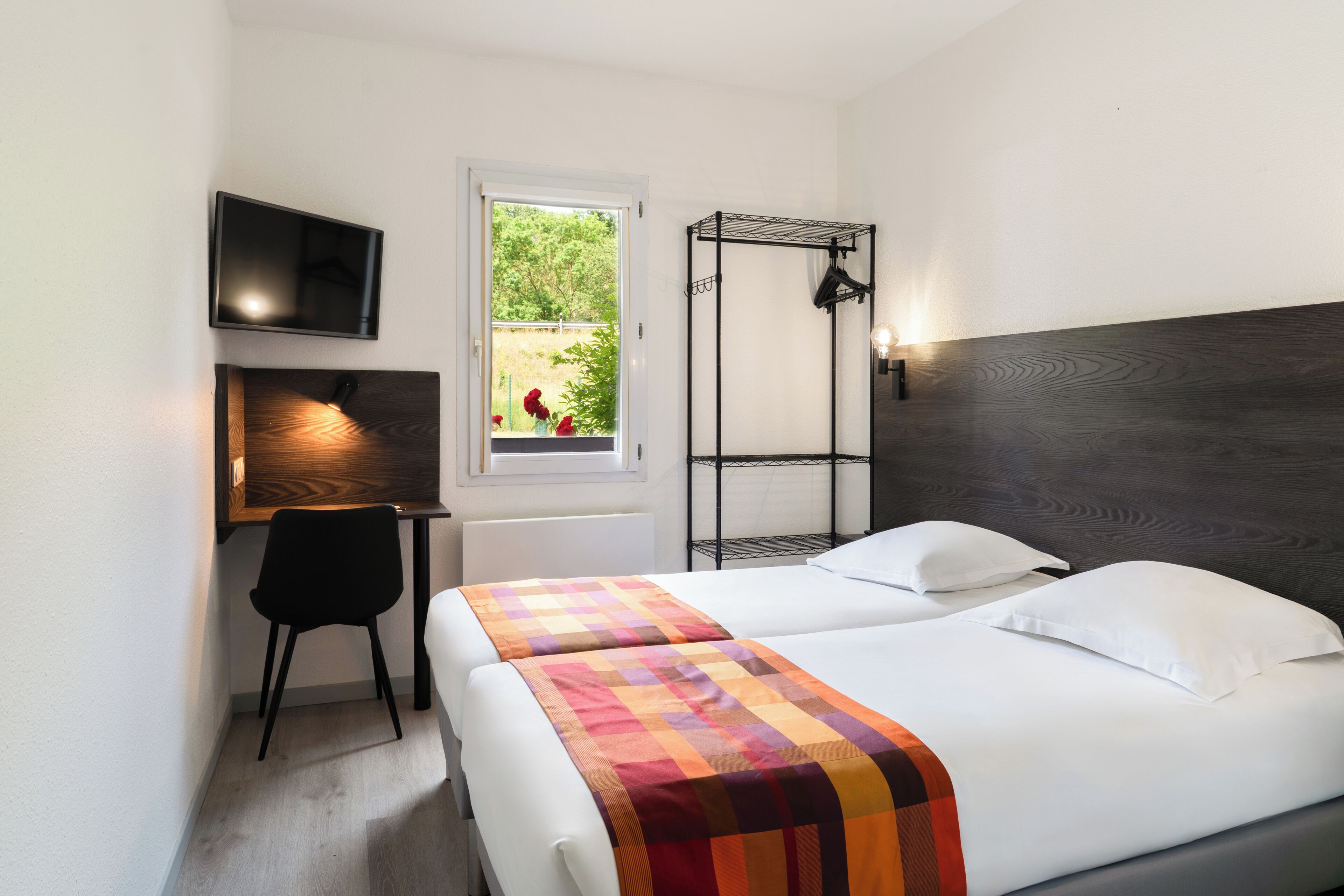 Een slaapkamer met een bed, een bureau met stoel, een aan de muur bevestigde televisie en een raam met uitzicht op groen.