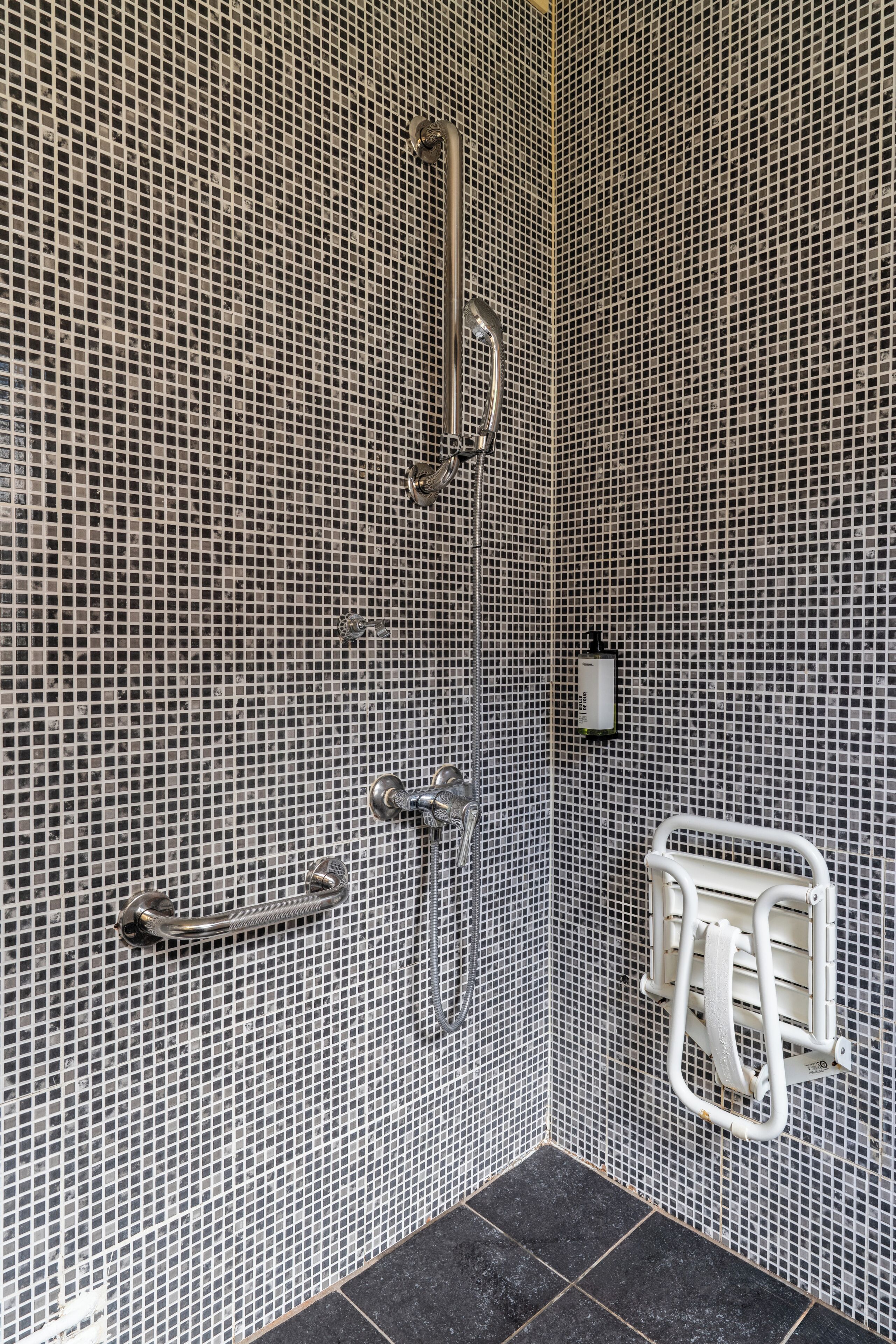 A bathroom with a tiled mosaic wall, a shower area, and a handrail.