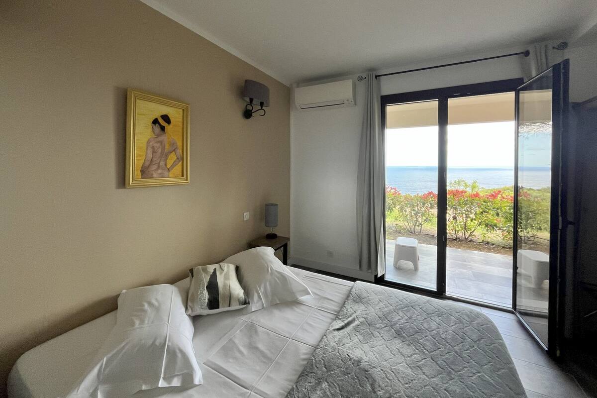 A bedroom with a bed, a painting on the wall, an air conditioning unit, and a view of the outdoors through a sliding glass door.