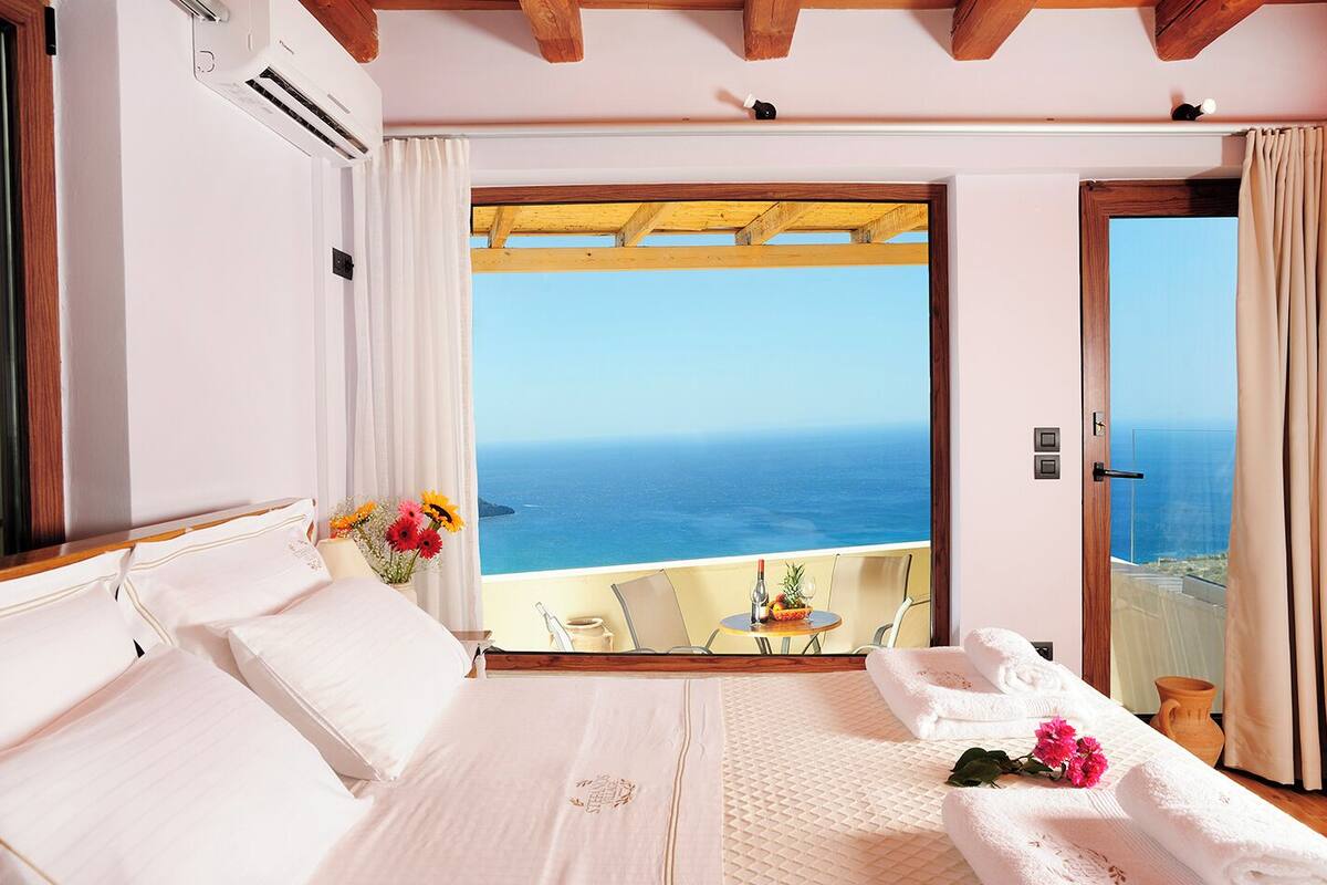 A bedroom with a large bed, a view of the sea, and a balcony with a table and chairs.