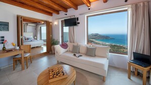 Un salon moderne avec un plafond en bois, un canapé blanc, une table basse en bois sur laquelle est posé un jeu d’échecs, un téléviseur fixé au mur et une grande fenêtre donnant sur la mer.