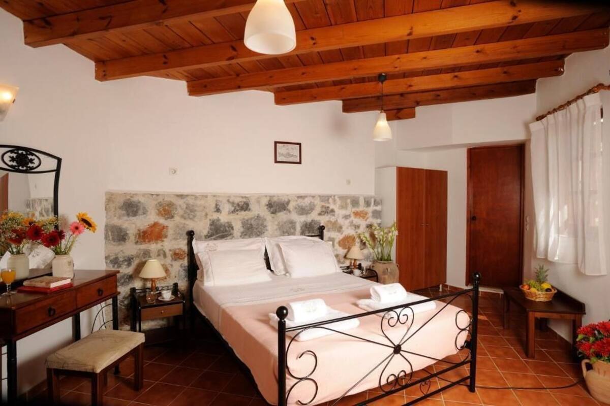 A bedroom with a stone wall, wooden ceiling beams, a metal bed frame, a bedside table with a lamp, and a window with curtains.