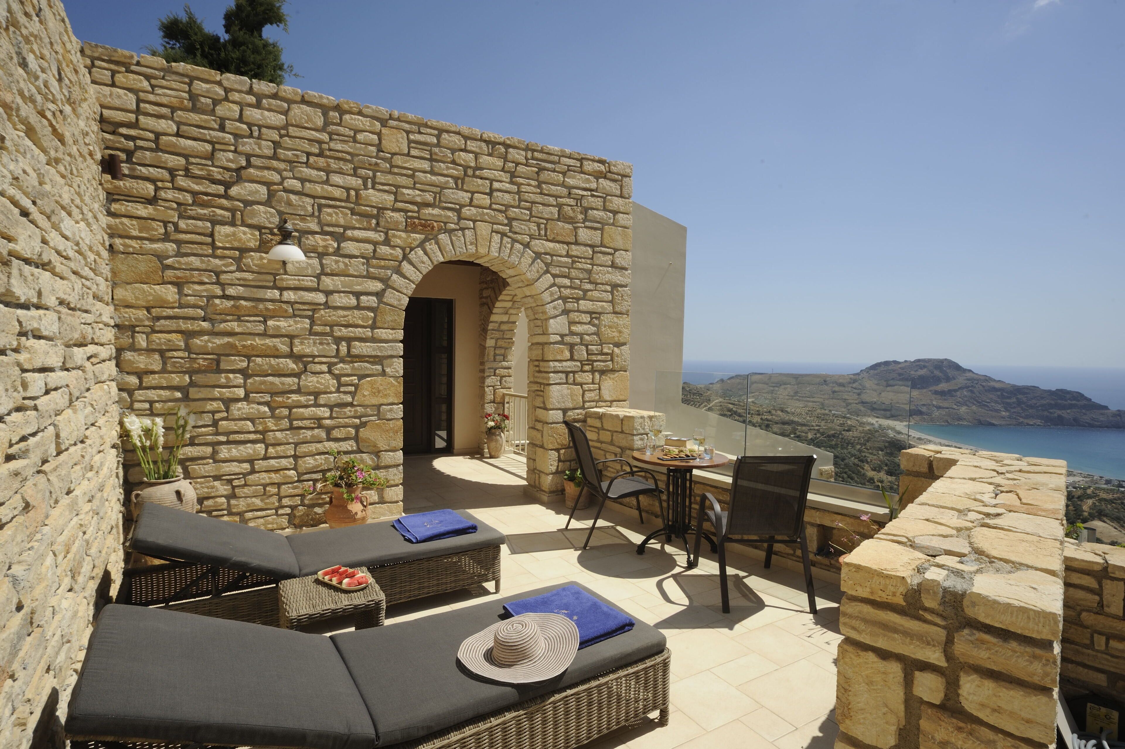 Un espace extérieur aménagé avec des murs en pierre, doté de mobilier en osier, d’une table et offrant une vue sur la mer et les collines.